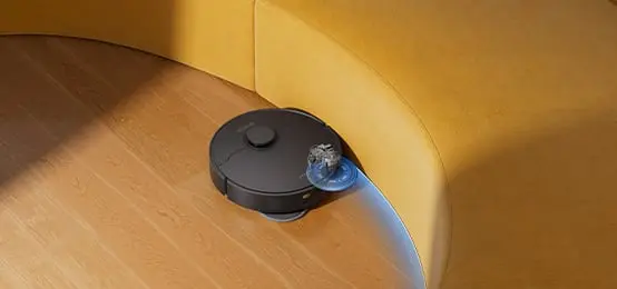 Melns robots putekļu sūcējs tīra grīdu gar dzeltenā dīvāna malu